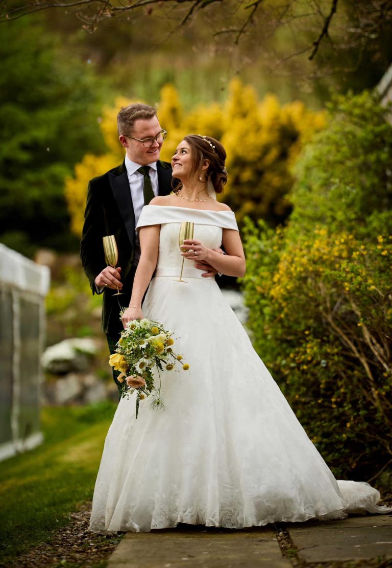 Couple together in garden, looking towards each bride holding flowers