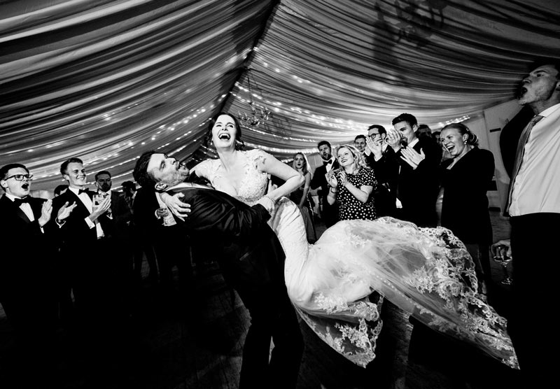 Black and white photography of the groom carrying the bride in a marquee on their wedding day for the best of 2025 Weddings
