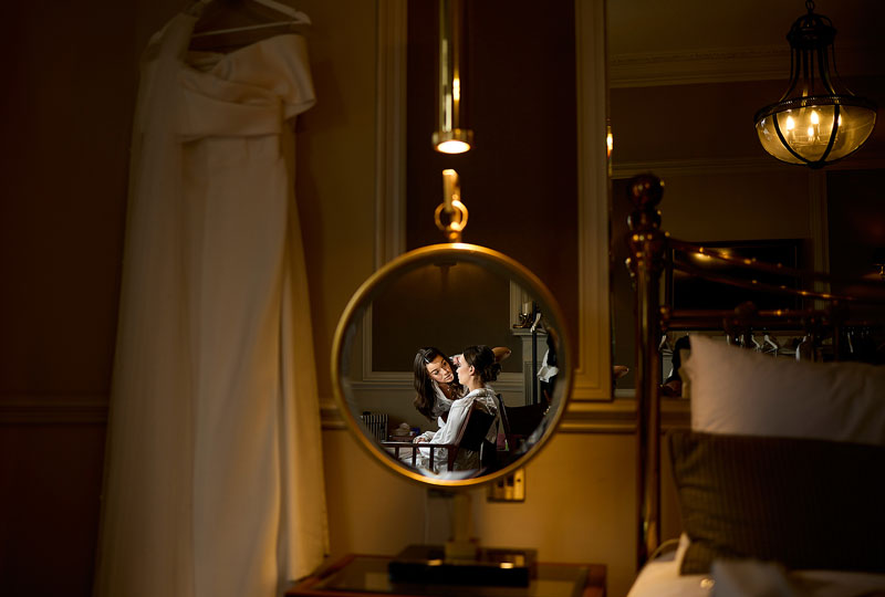 reflection of a Bride having her make-up done in a hanging mirror with the wedding dress in the background for the discussion on Choosing a wedding photographer